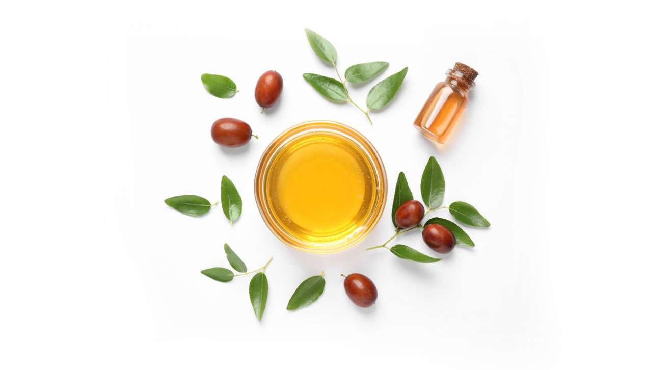 Small bowl of jojoba oil surrounded by several leaves and beans